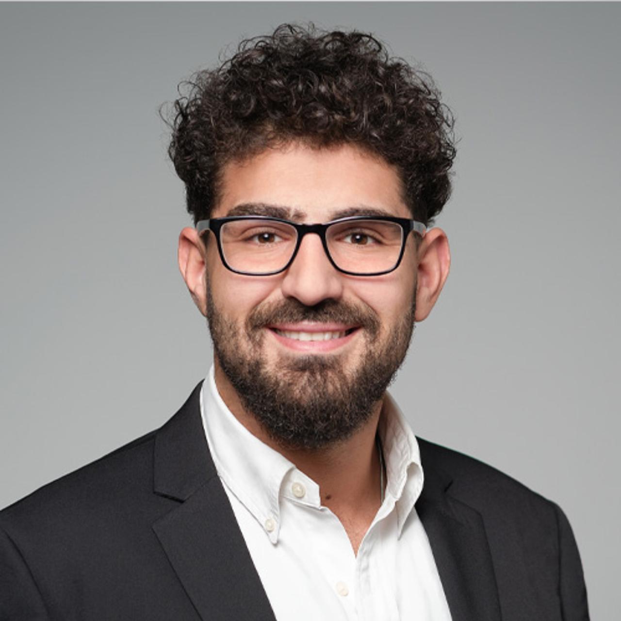 Person_mit_lockigem_Haar_und_Bart_trägt_ein_weisses_Hemd_und_schwarzen_Blazer